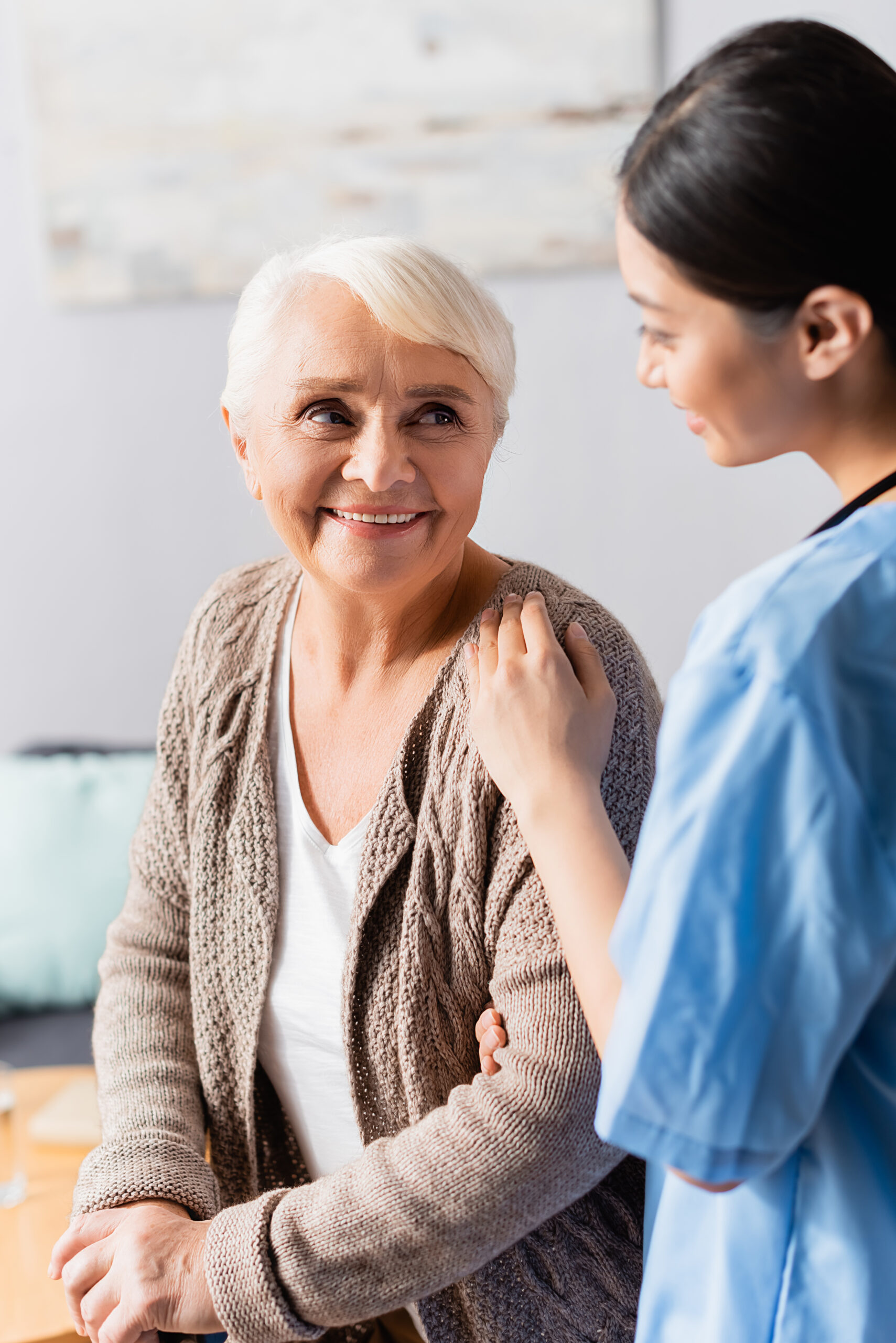 young asian nurse touching shoulder of smiling senior woman in nursing home, blurred foreground